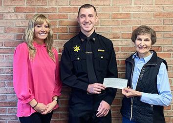 A woman stands to the left of a man in a police officer's uniform. They are holding a check and there is a woman standing with them to the right smiling with her hands folded.